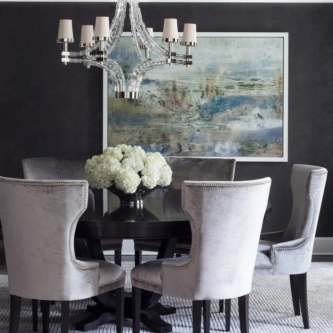 Pale finely striped rug beneath a dark round table and gray upholstered chairs against a dark wall