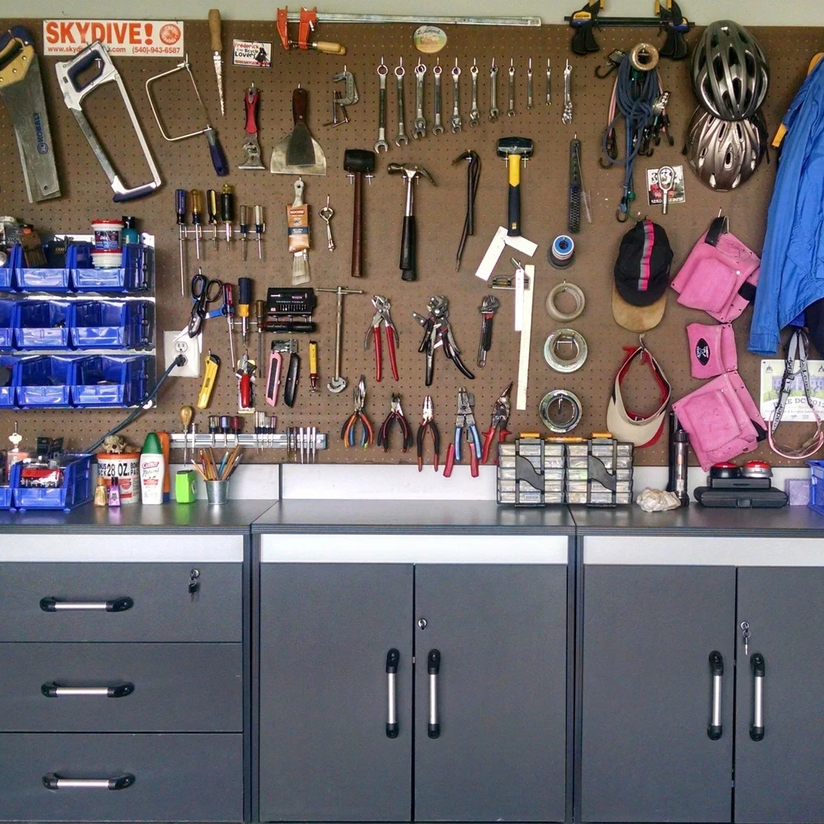 Garage workbench with brown pegboard, rows of hand tools, blue parts bins and dark lower cabinets.