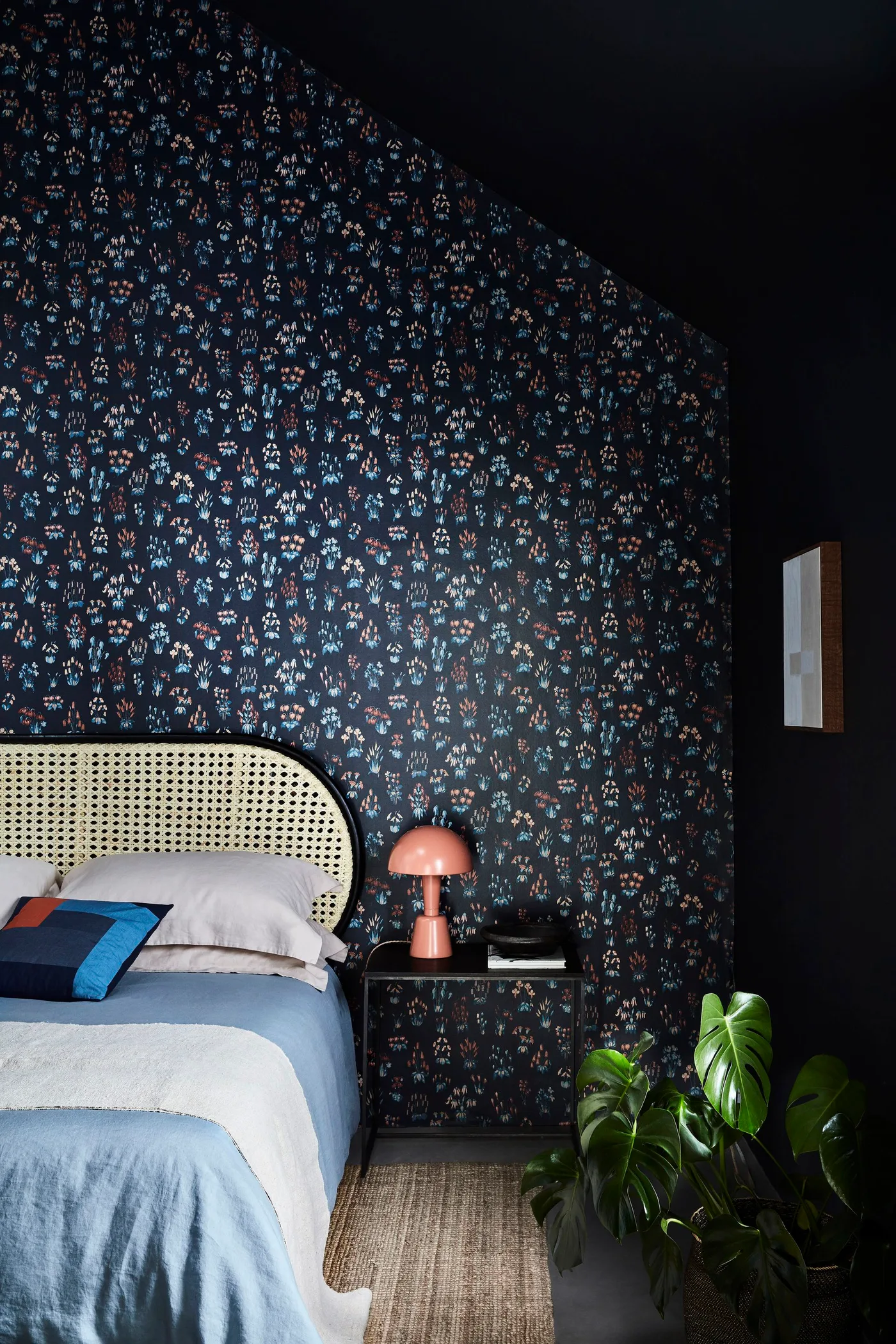 Small multicolored flowers on dark wallpaper above a cane headboard in a sloped-ceiling bedroom.