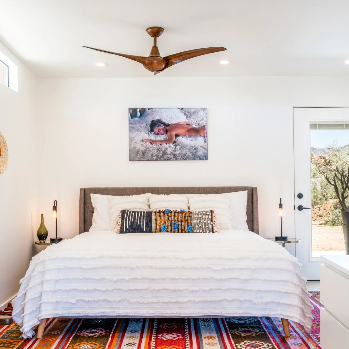 White bedroom with a simple upholstered bed, colorful geometric rug, narrow bedside tables and a long textured cushion
