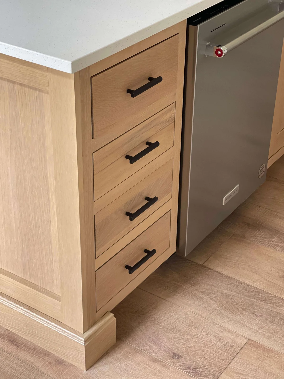 Inset oak drawers with black pulls sit beside a stainless dishwasher below a pale speckled countertop and an oak island end.