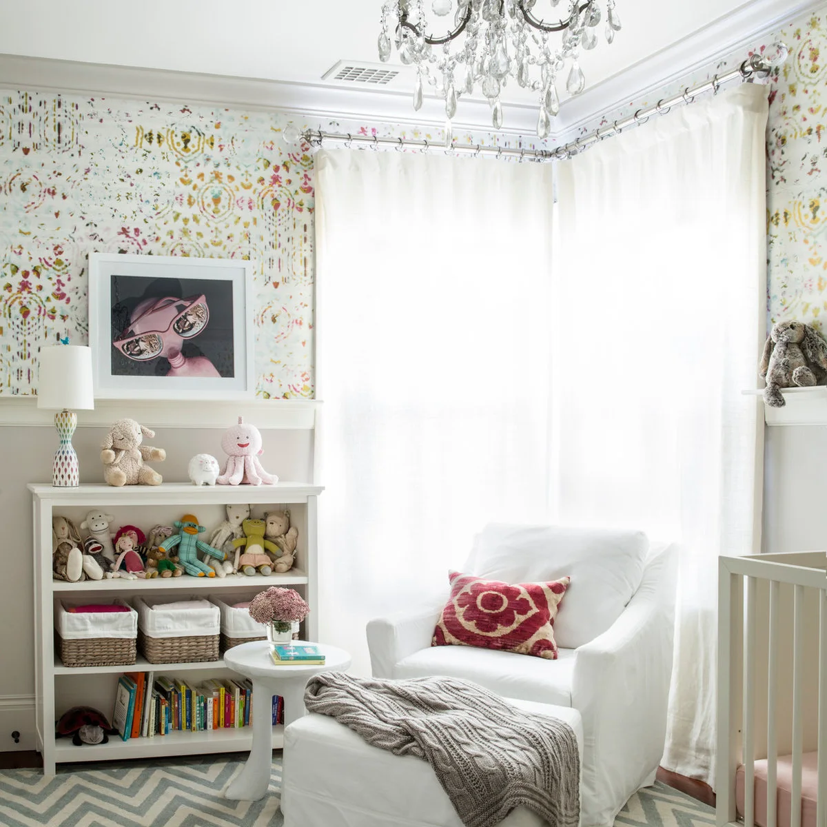 White nursery bookcase with toys, lined baskets and books beside a pale armchair and ottoman