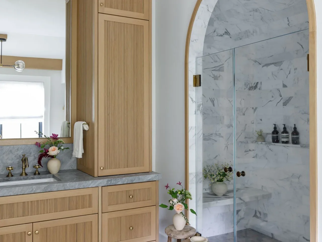 Pale ribbed timber cabinet fronts sit beside an arched shower opening with pale veined tiles.