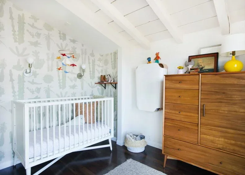 White nursery crib beside tall wood dresser, pale illustrated wallpaper, white board ceiling and yellow lamp