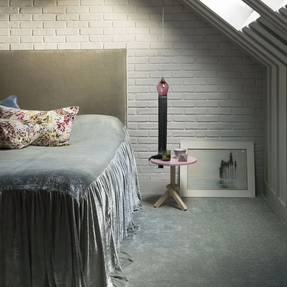 Small pink bedside table below a hanging pink light beside an upholstered bed in a white brick attic room.