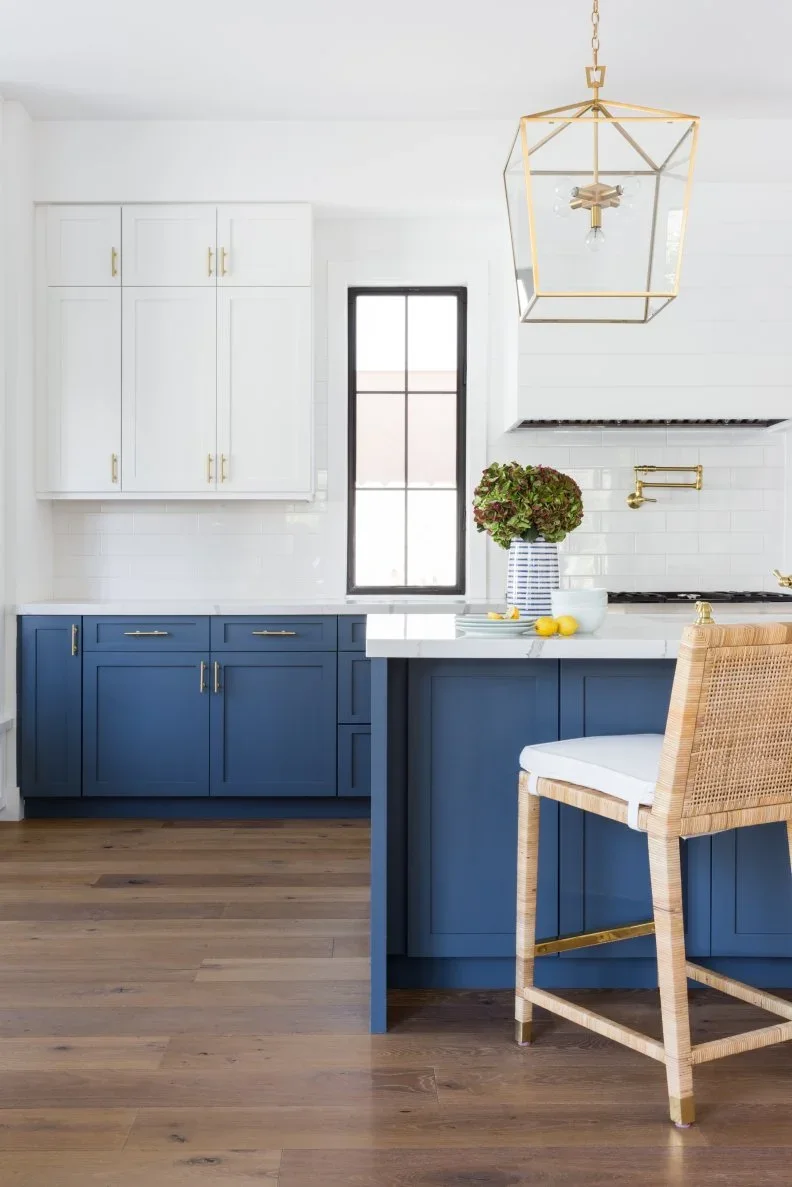 Blue lower cabinets and an island contrast with white uppers and a white hood, with a gold-colored lantern and a woven stool.