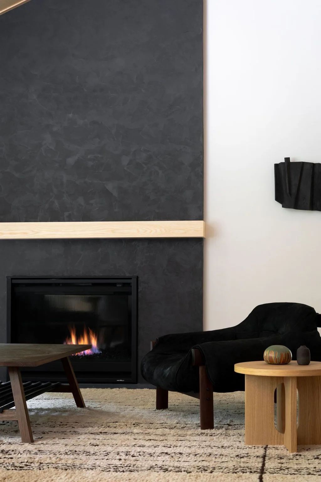 Pale living room corner with a black lounge chair, low wooden side table, textured rug and dark fireplace surround.
