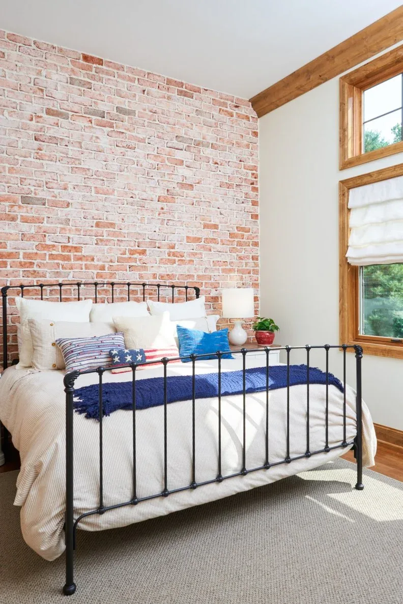 A black metal bed stands against a pale brick wall beside a tall timber-trimmed window and white bedding.