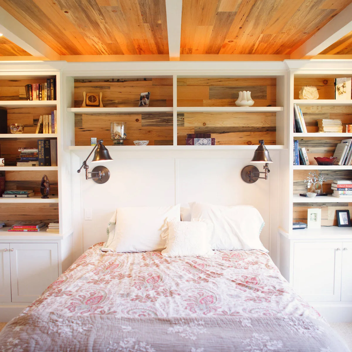 A bed between white shelving units with wood backing and separate adjustable wall lights above each pillow.