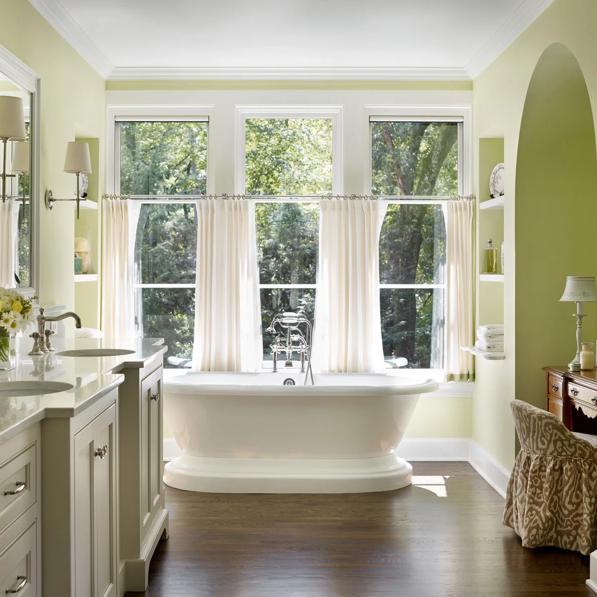 Cream cafe curtains cover lower portions of three tall windows behind a freestanding bath in a pale green bathroom.