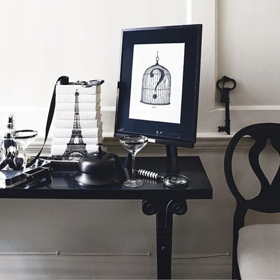 A small glossy black table and dark curved-back chair sit beside pale walls with a black-framed print, stacked books and clear glasses