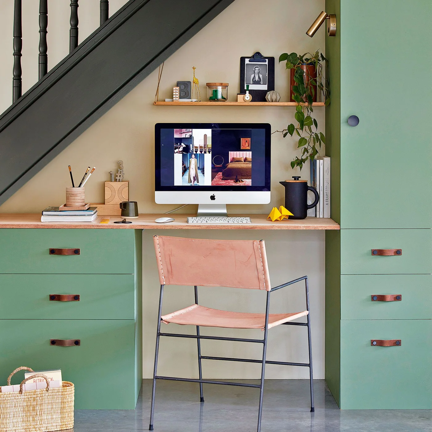 Green drawer units flank a desk beneath a staircase, with a pink chair, a narrow shelf, a monitor and a small wall light.