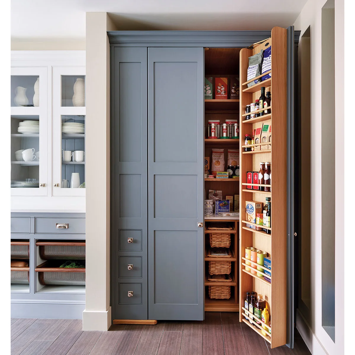 A blue-gray larder cabinet opens to show wood shelves, lower baskets and shallow bottle racks mounted inside the door.