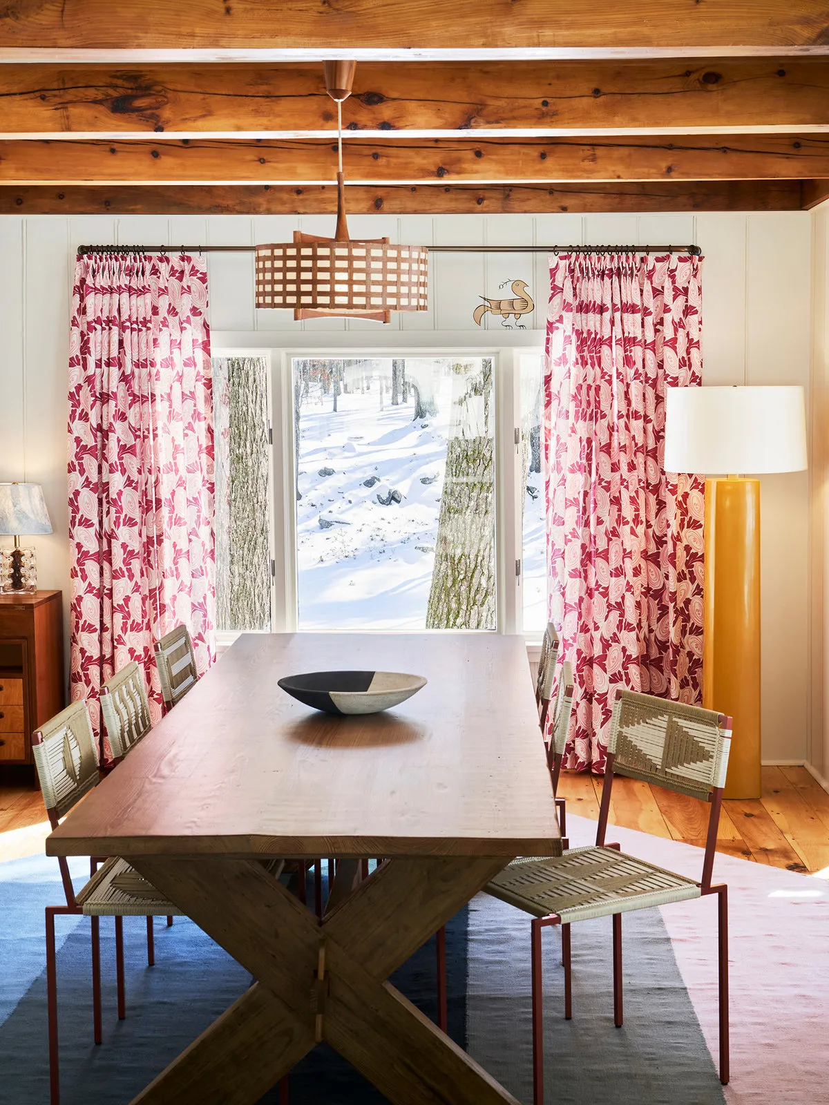 Pink patterned curtains frame a timber dining table beneath an exposed wood ceiling.