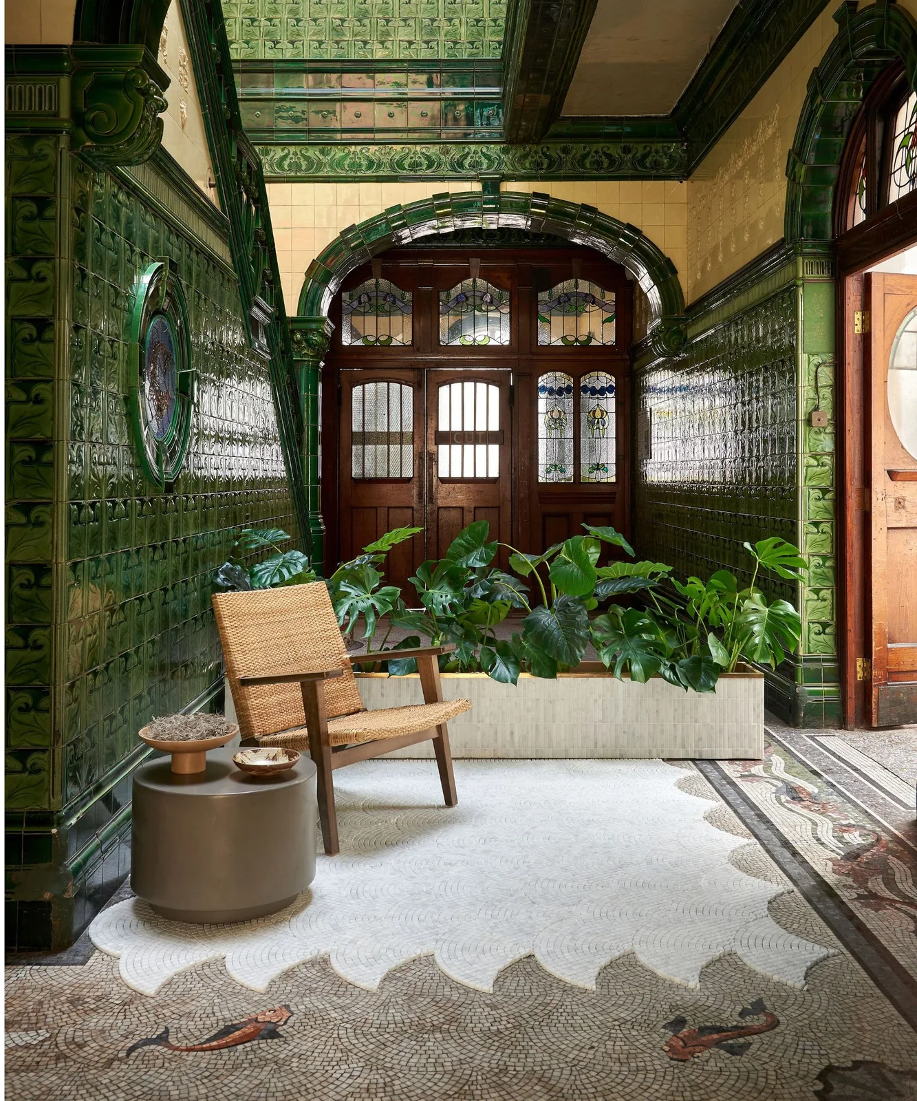 A fish-and-wave mosaic floor runs through a green tiled entry hall below a woven chair and broad planter.