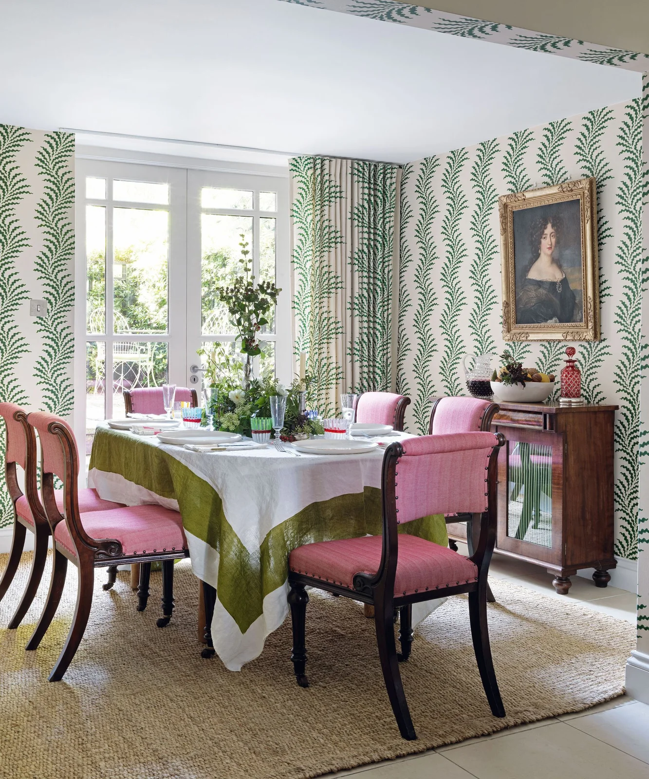 Green frond-patterned walls and curtains surround a pale-clothed dining table with pink upholstered chairs and a wooden cabinet