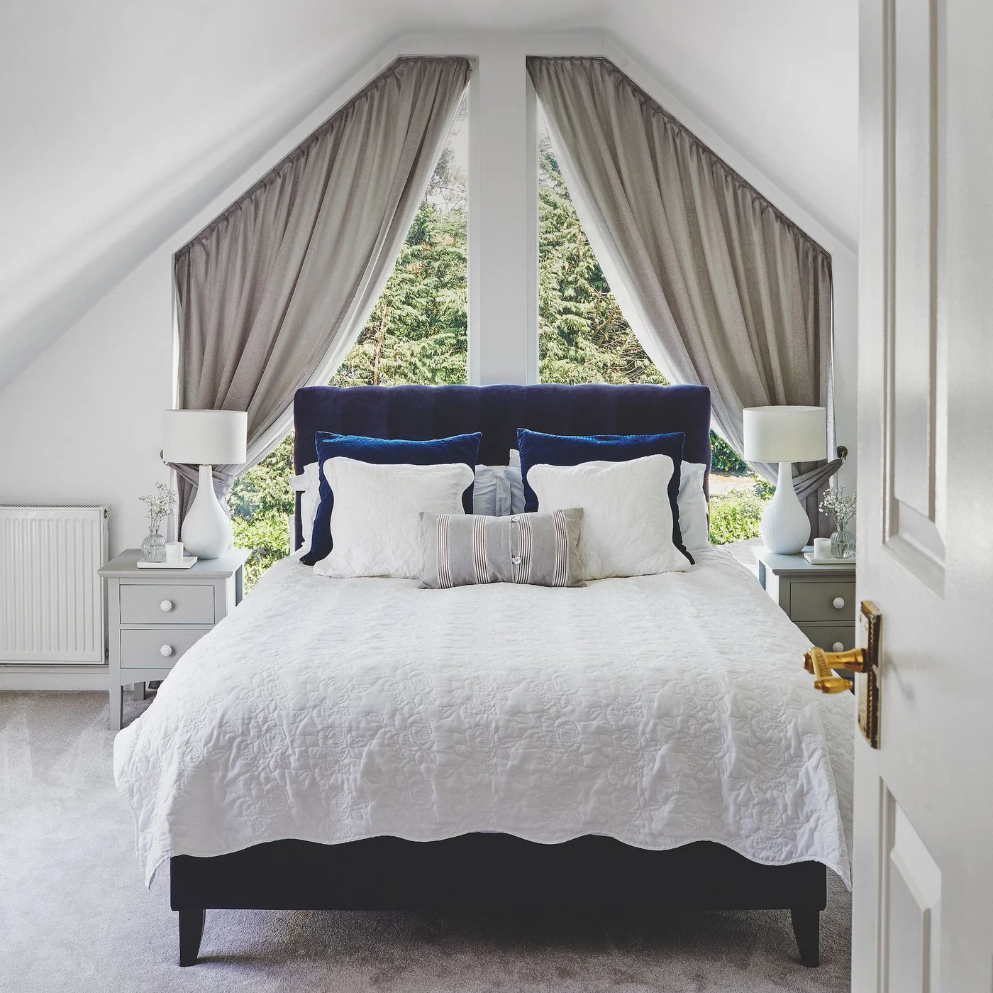 Blue headboard below a shaped gable window with gray curtains and white bedding in a sloped-ceiling bedroom.