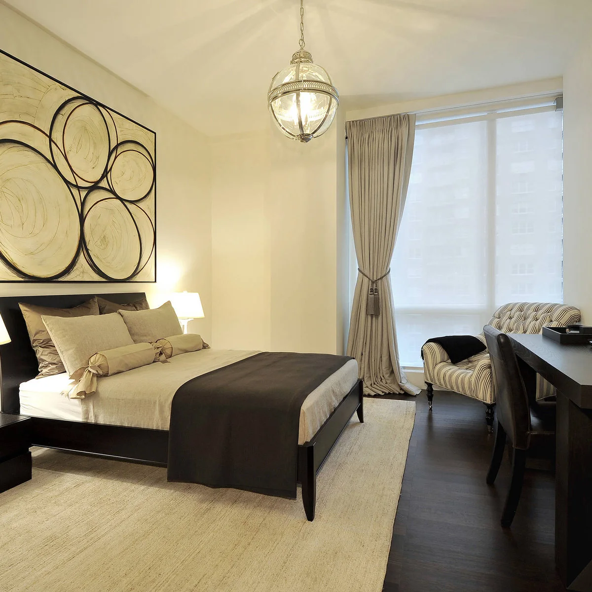 Bedroom with a dark wood bed, narrow desk, large circular artwork and a striped chair beside the window