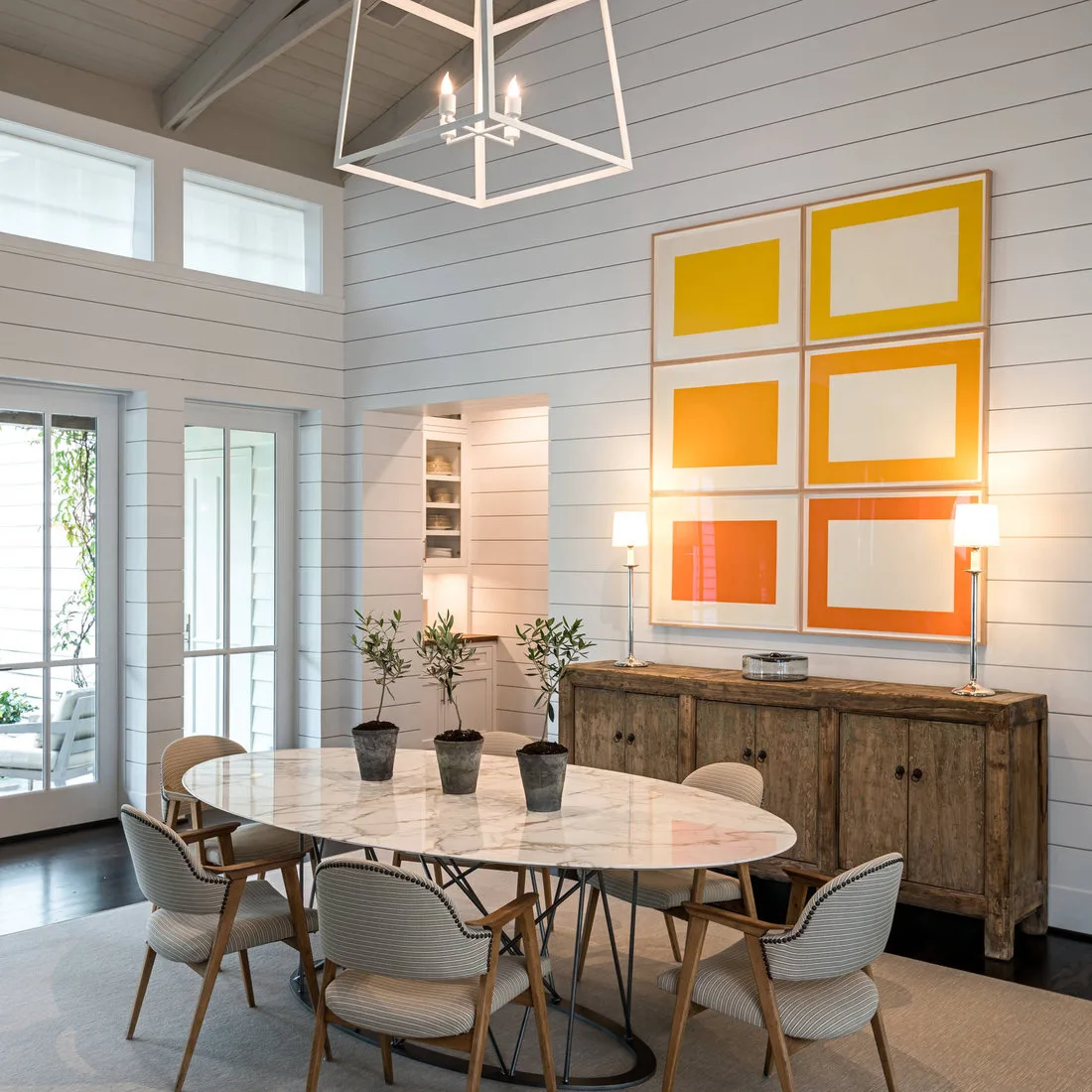 Pale oval dining table with patterned chairs beside a wood sideboard and six bright framed artworks