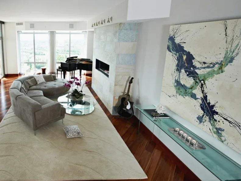 Living room with curved gray sectional, round glass table, pale rug, large abstract artwork, low glass-fronted console and piano beside broad windows.