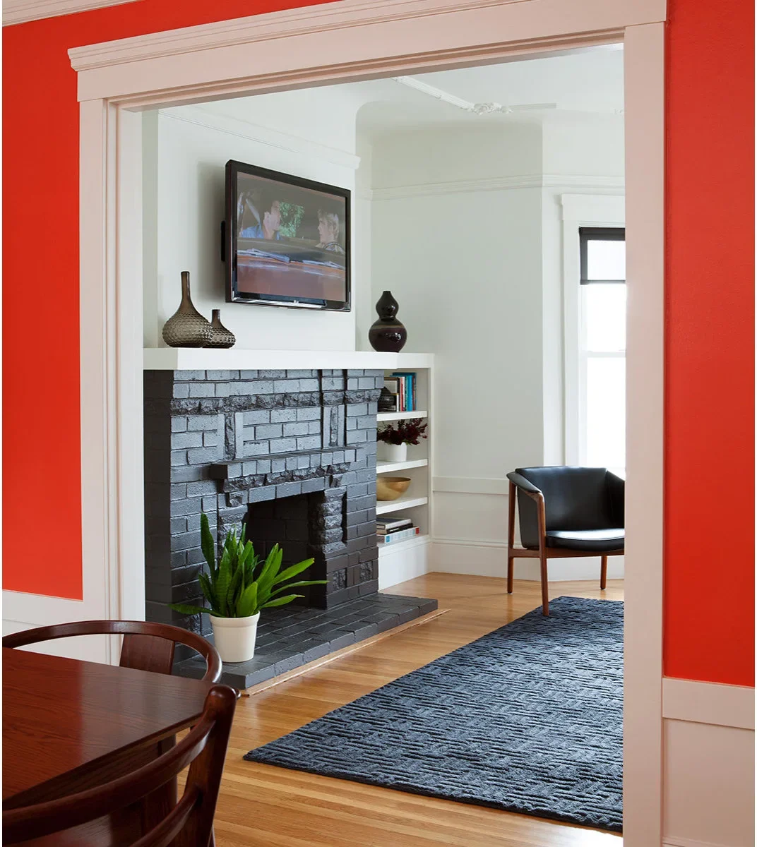 A charcoal-painted brick fireplace with a pale shelf sits in a white room beyond a red-painted doorway.