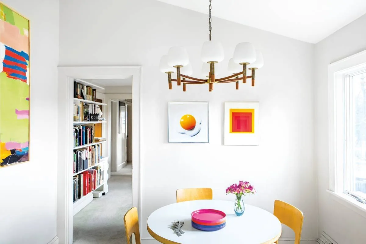 Two bright pictures in white frames above yellow chairs and a white round dining table