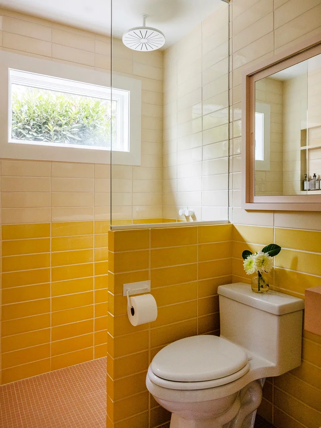 Cream and mustard bathroom tiles form two horizontal bands beside a white toilet, white-framed window and wood-framed mirror with a round showerhead above