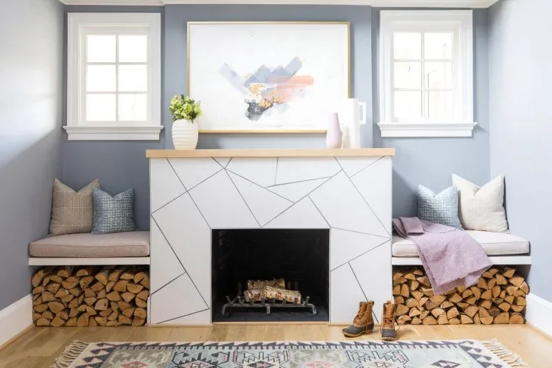 White geometric fireplace surround with wood mantel between two windows, cushioned benches and stored logs