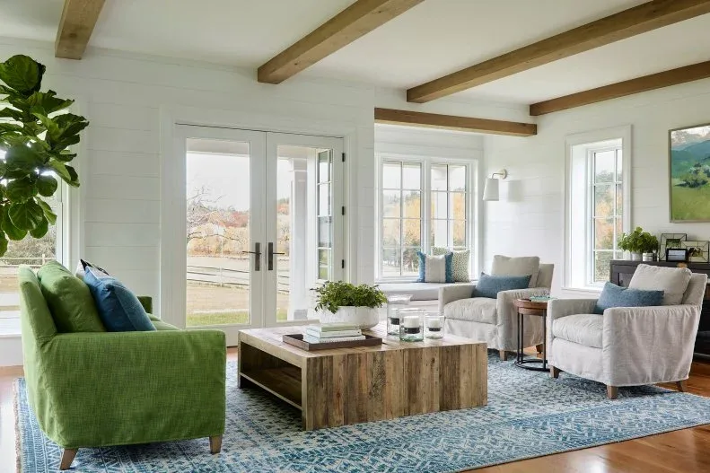 A green sofa and pale armchairs face a broad wooden coffee table beneath wooden beams above a blue patterned rug