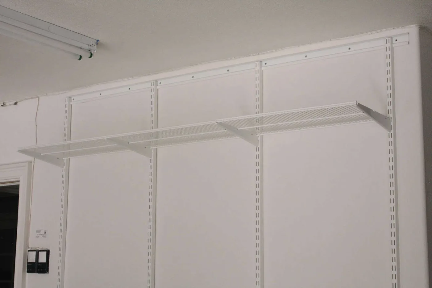 White top rail and vertical shelf supports with one long open shelf on a pale garage wall.