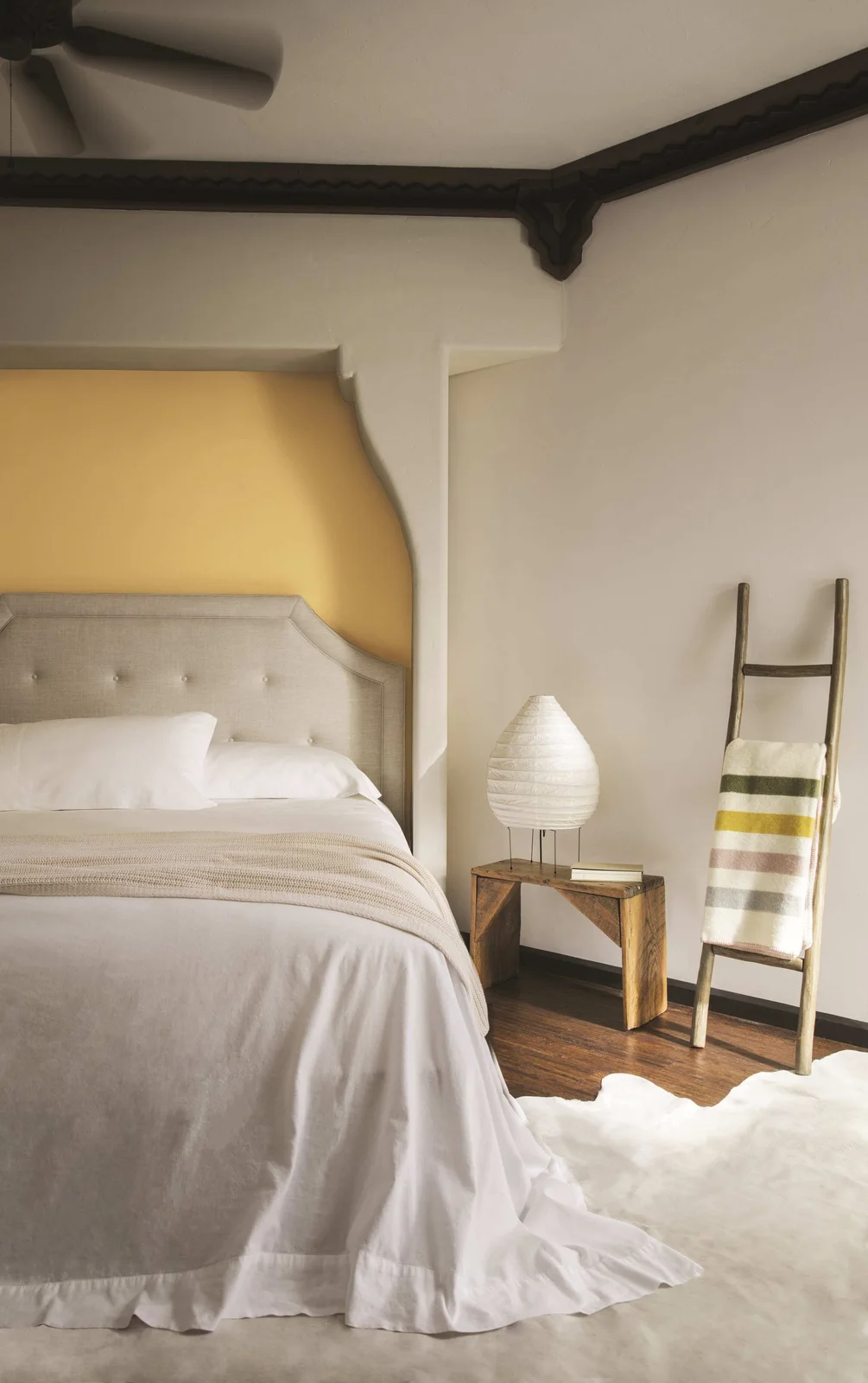 A muted mustard recess frames a neutral upholstered headboard beside a block-shaped wood table, a pale lamp and a blanket ladder.