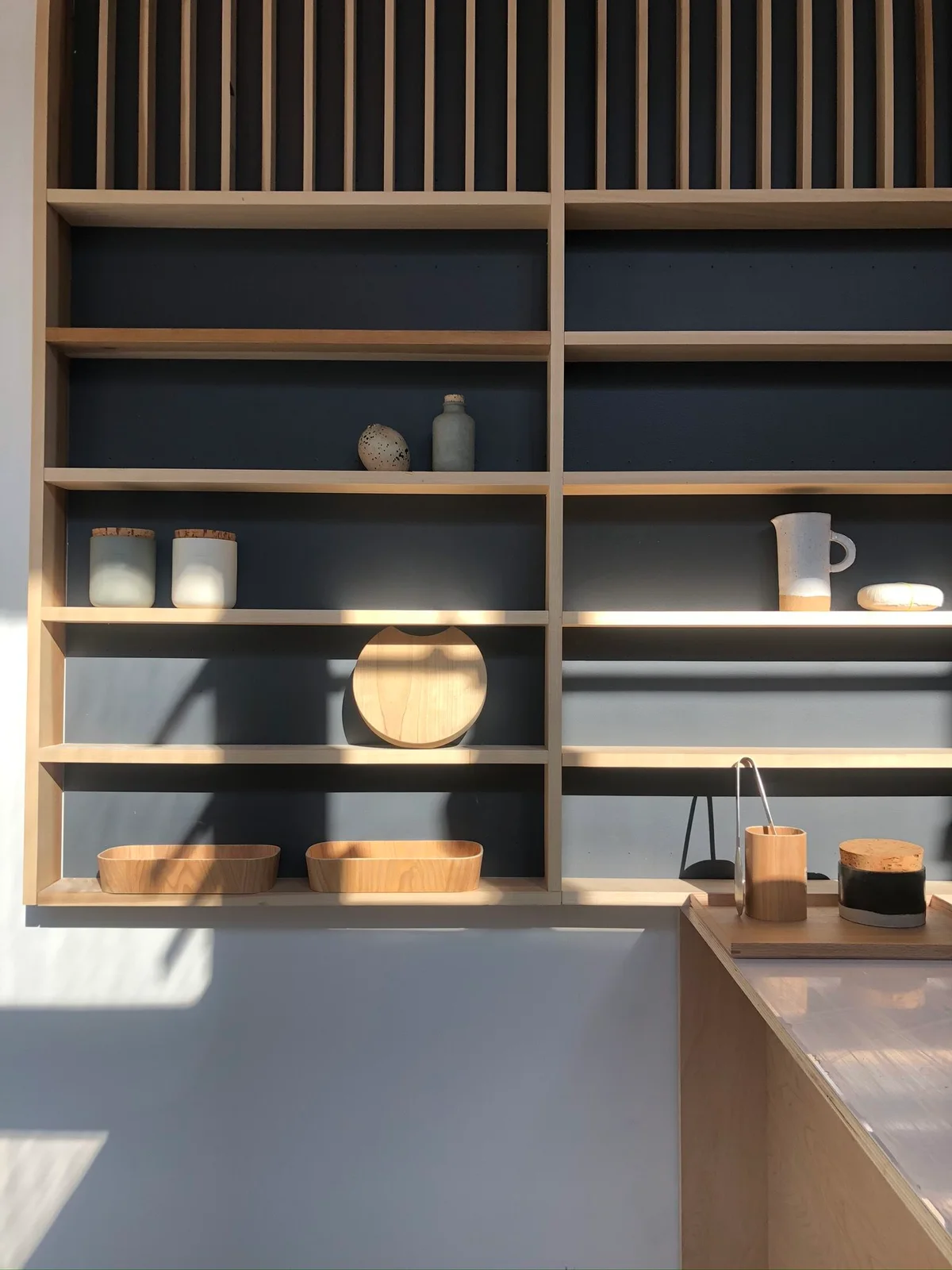 A pale wooden grid shelf holds a few bowls, jars and vessels against a dark backing beside a light counter