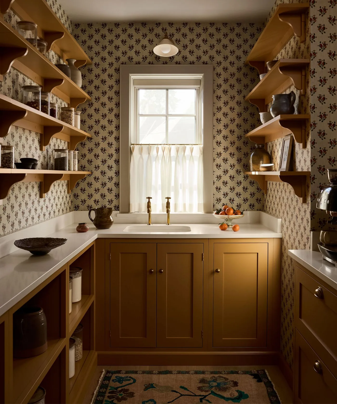 Country pantry with ochre shelves and cabinets, floral wallpaper, pale sink and cafe-curtained window
