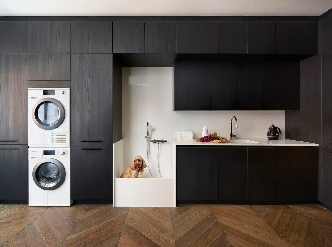 Dark cabinetry surrounds stacked laundry machines, a low dog-washing recess and a sink counter above a wood-patterned floor.