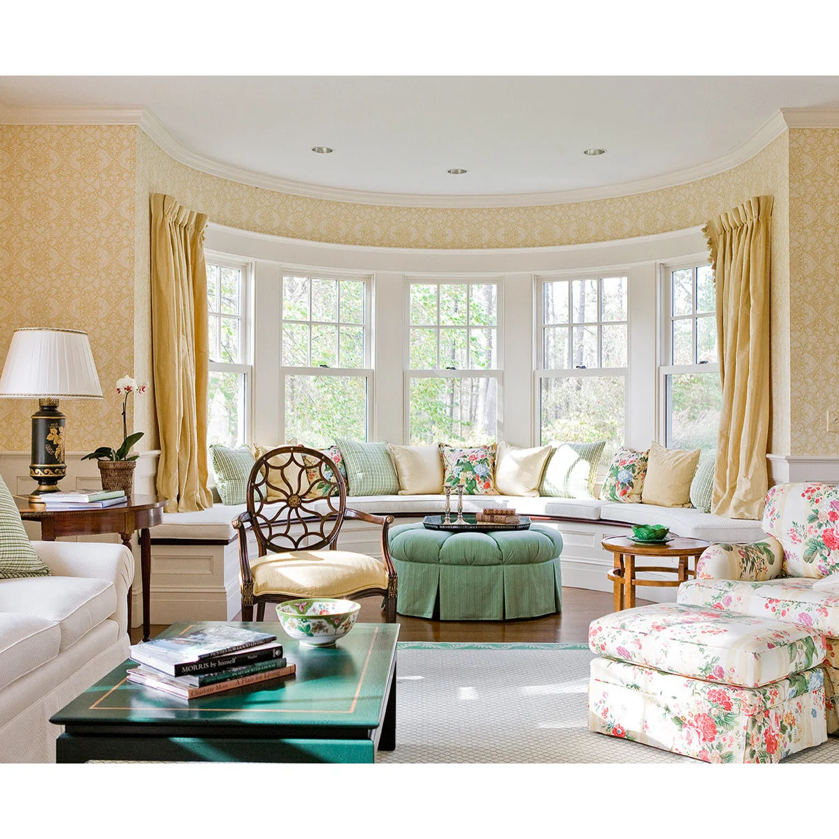 A curved window seat with pale green and floral cushions in a yellow living room with a round green ottoman.