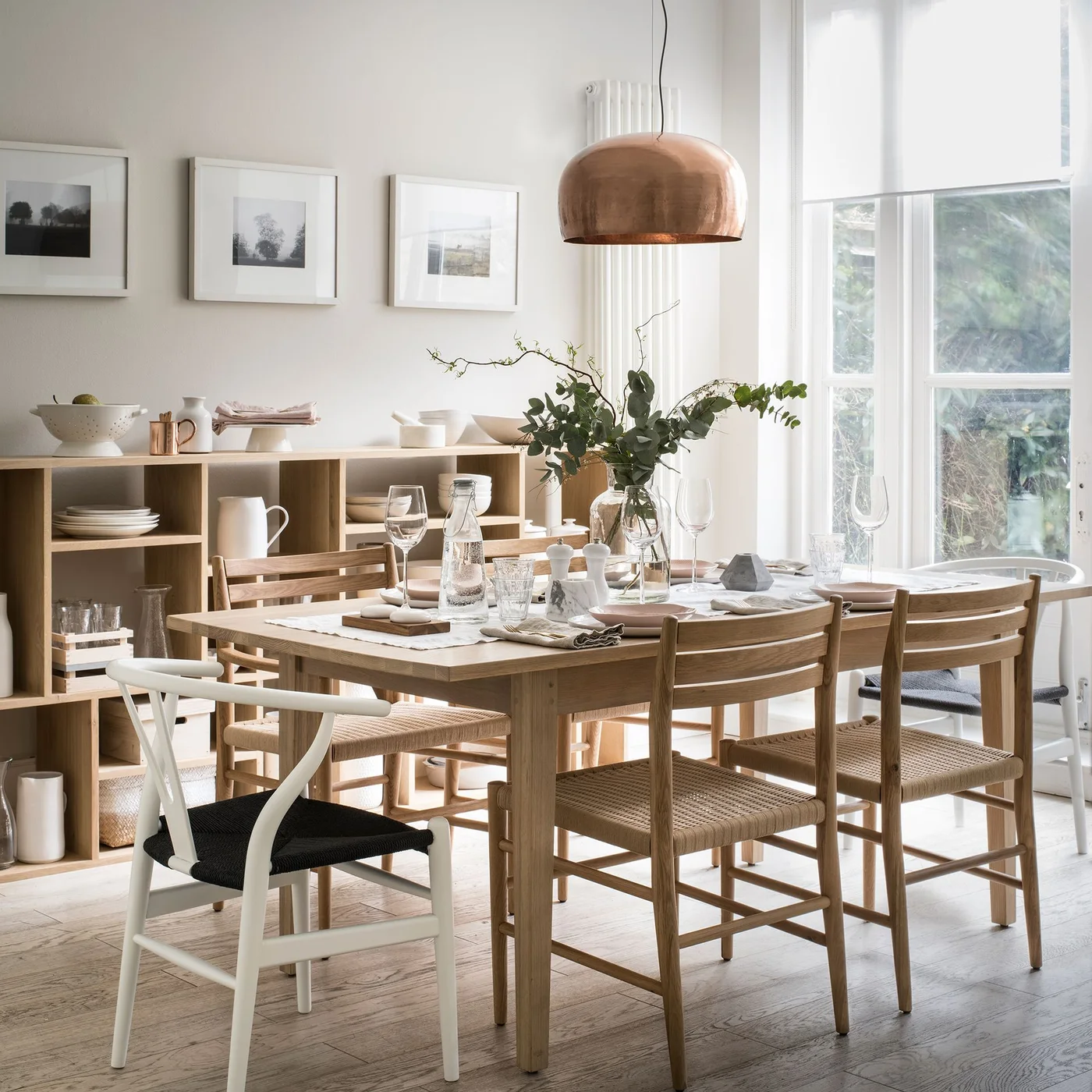 Low open wood shelves hold plates, cups and bowls beside a wood dining table with woven-seat chairs and a pale wall