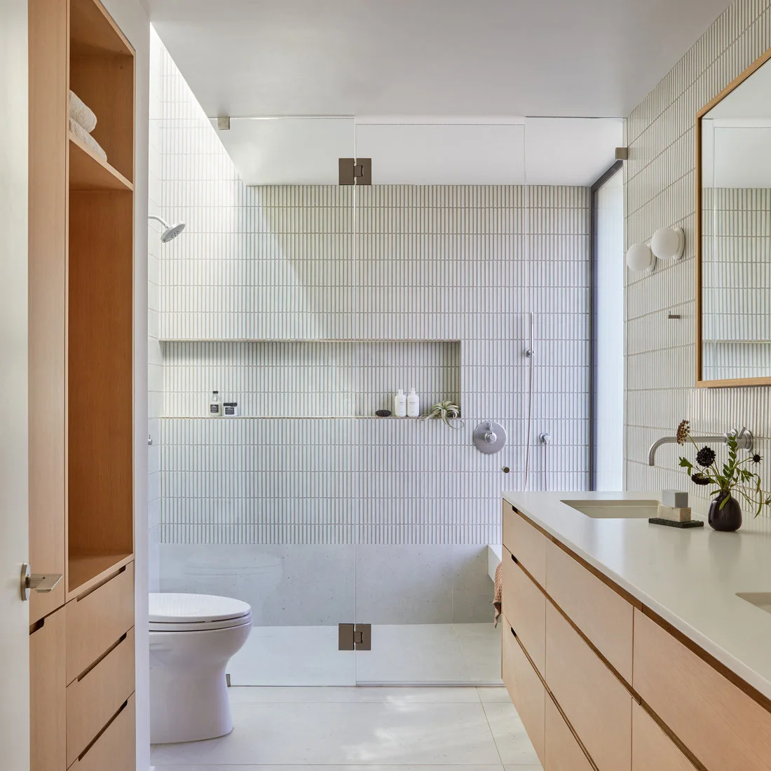 Slender pale vertical shower tiles surround a broad niche beside wooden cabinetry and a simple glass door