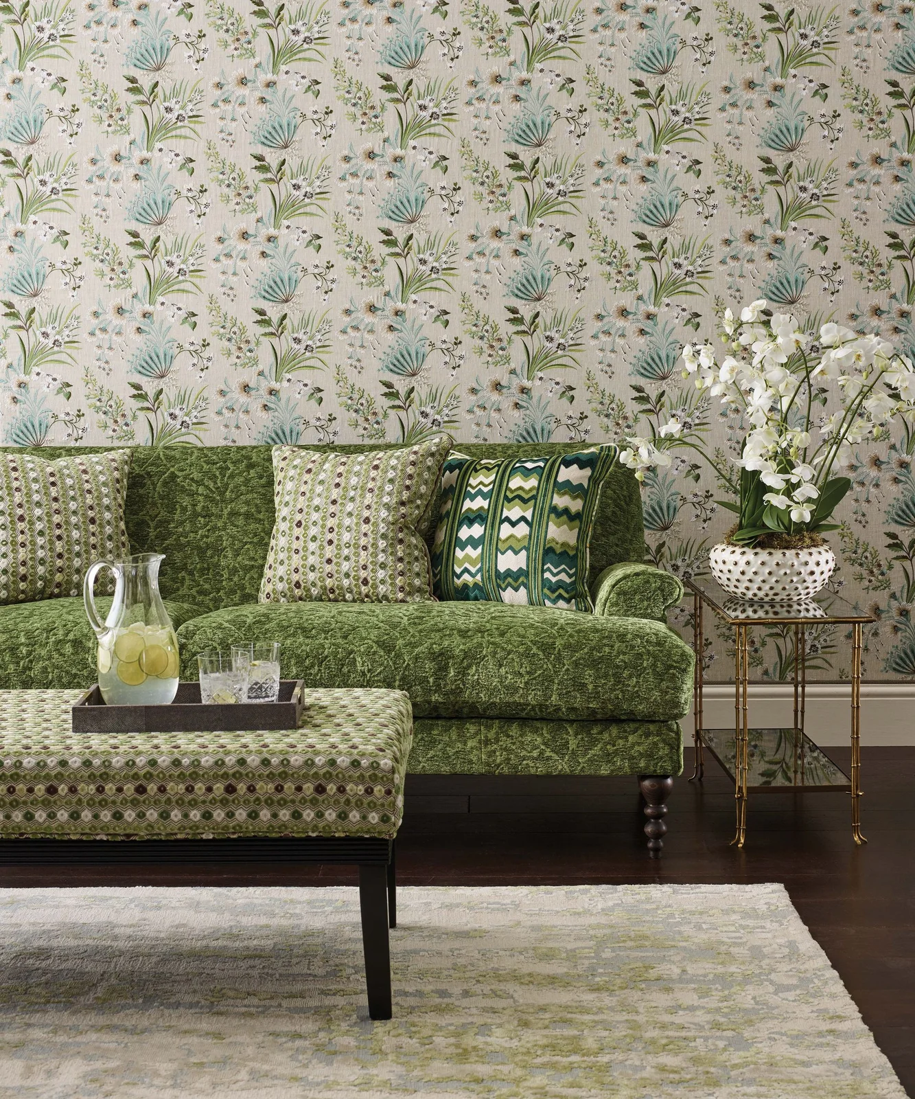 A patterned green sofa and dotted green footstool sit below botanical wallpaper beside a glass-topped side table