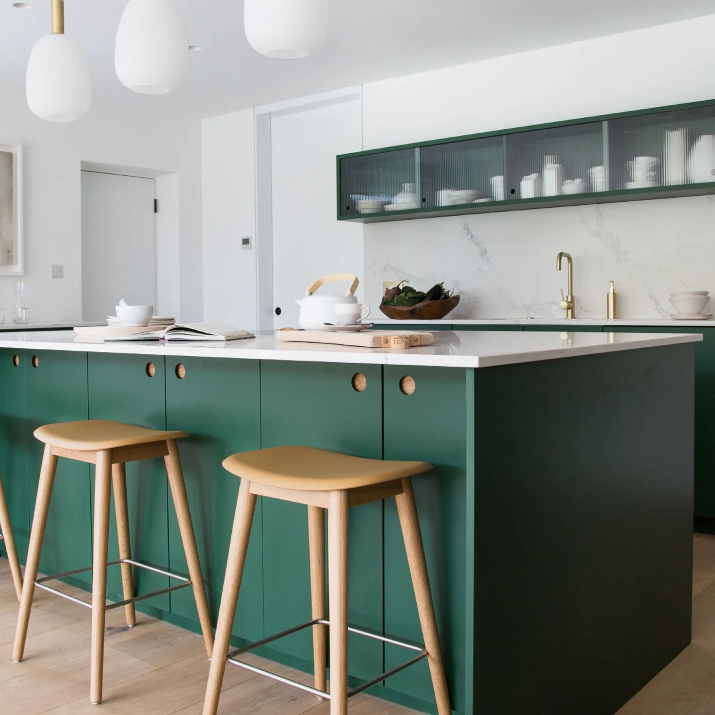 Deep green island fronts and a narrow glass-fronted upper cabinet sit against a light wall beside wooden stools