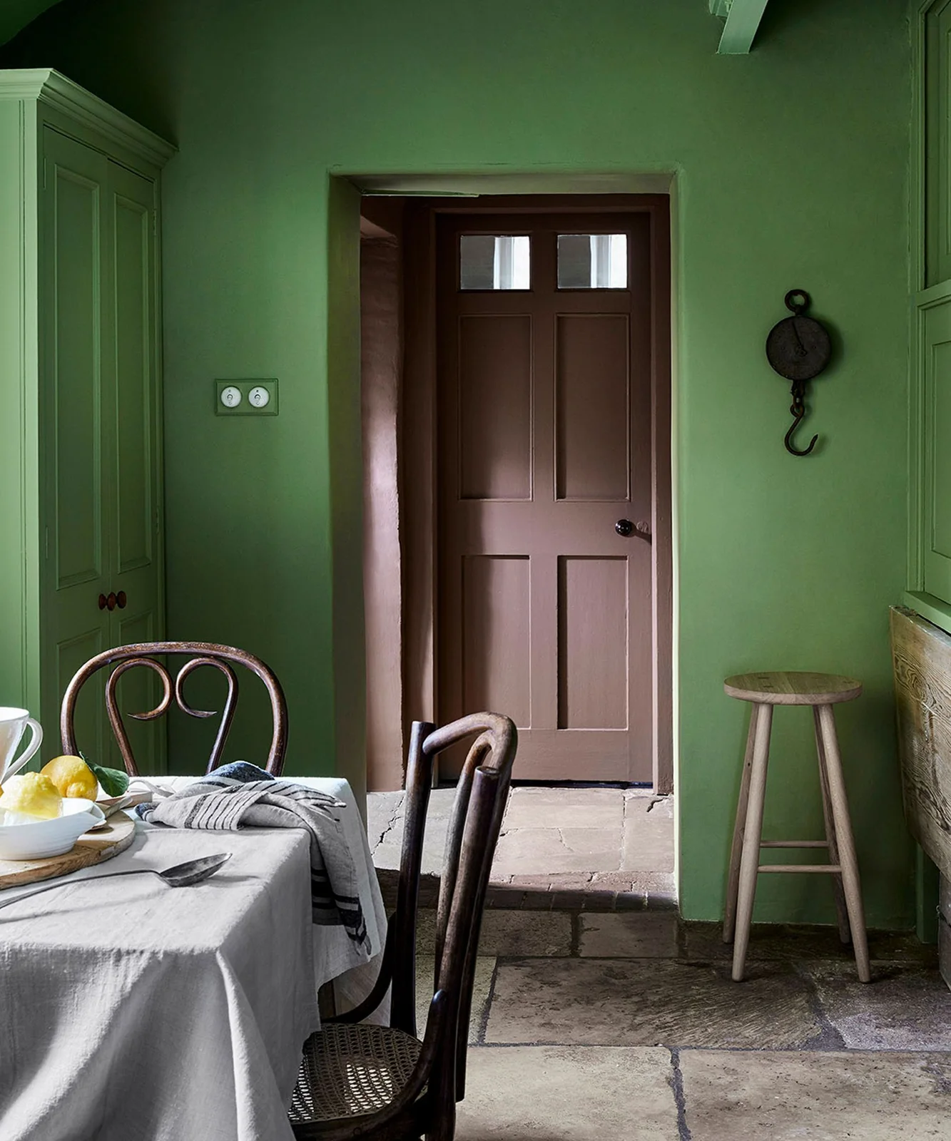 Green walls and painted storage stand beside a brown door, simple wooden stool and dining table with dark curved chairs and a pale cloth