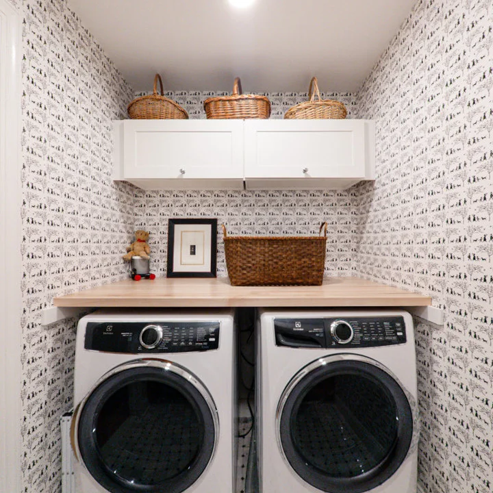 White cupboards and a wood counter surround two laundry machines beneath a fine black-and-white patterned backsplash and woven baskets.