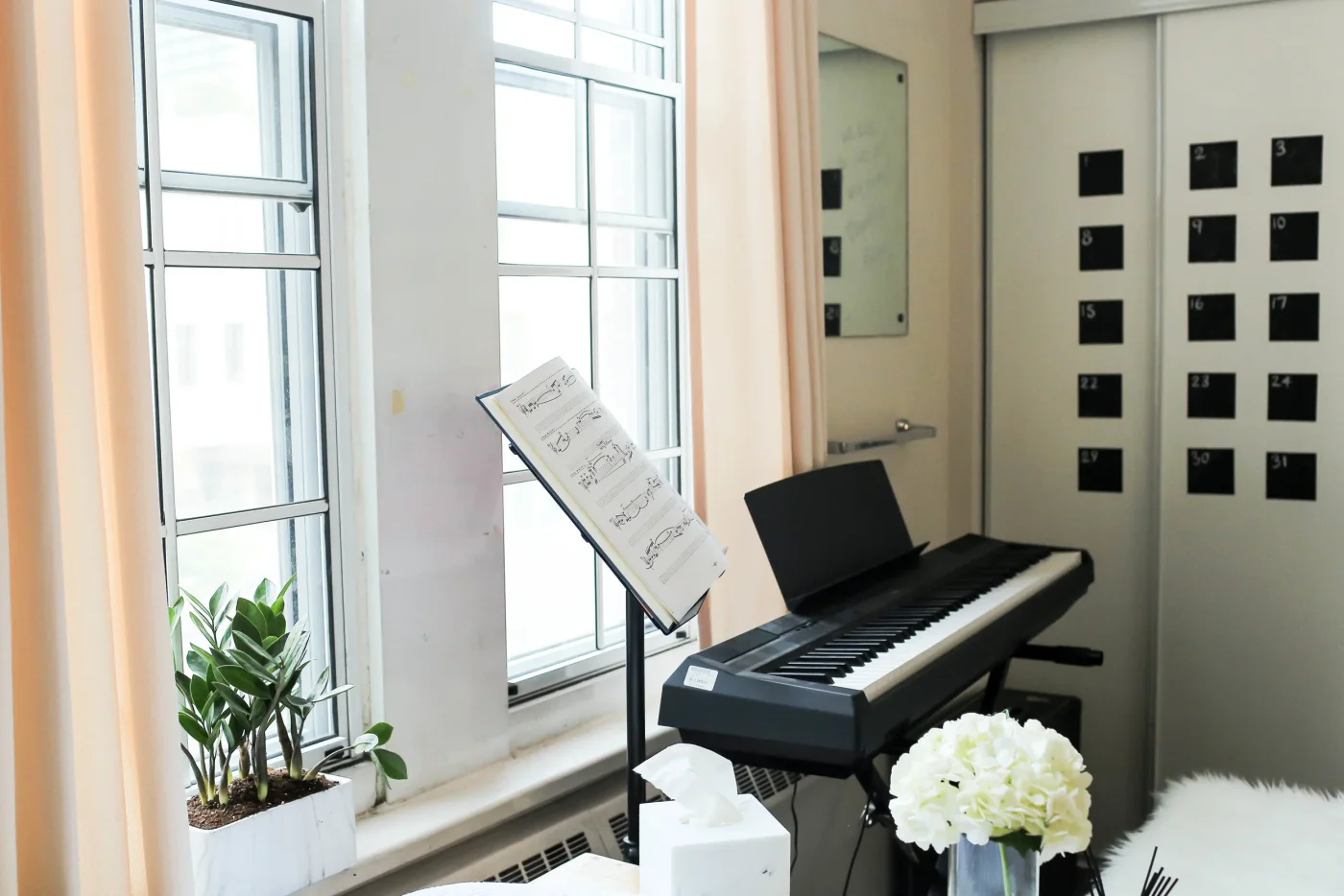 Keyboard and music stand beside tall dorm windows with a small plant on the sill and closet doors beyond