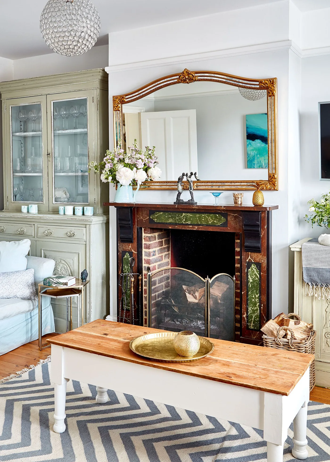 Decorative red-and-green fireplace beneath gold-framed mirror beside painted cupboard and white table with wooden top.