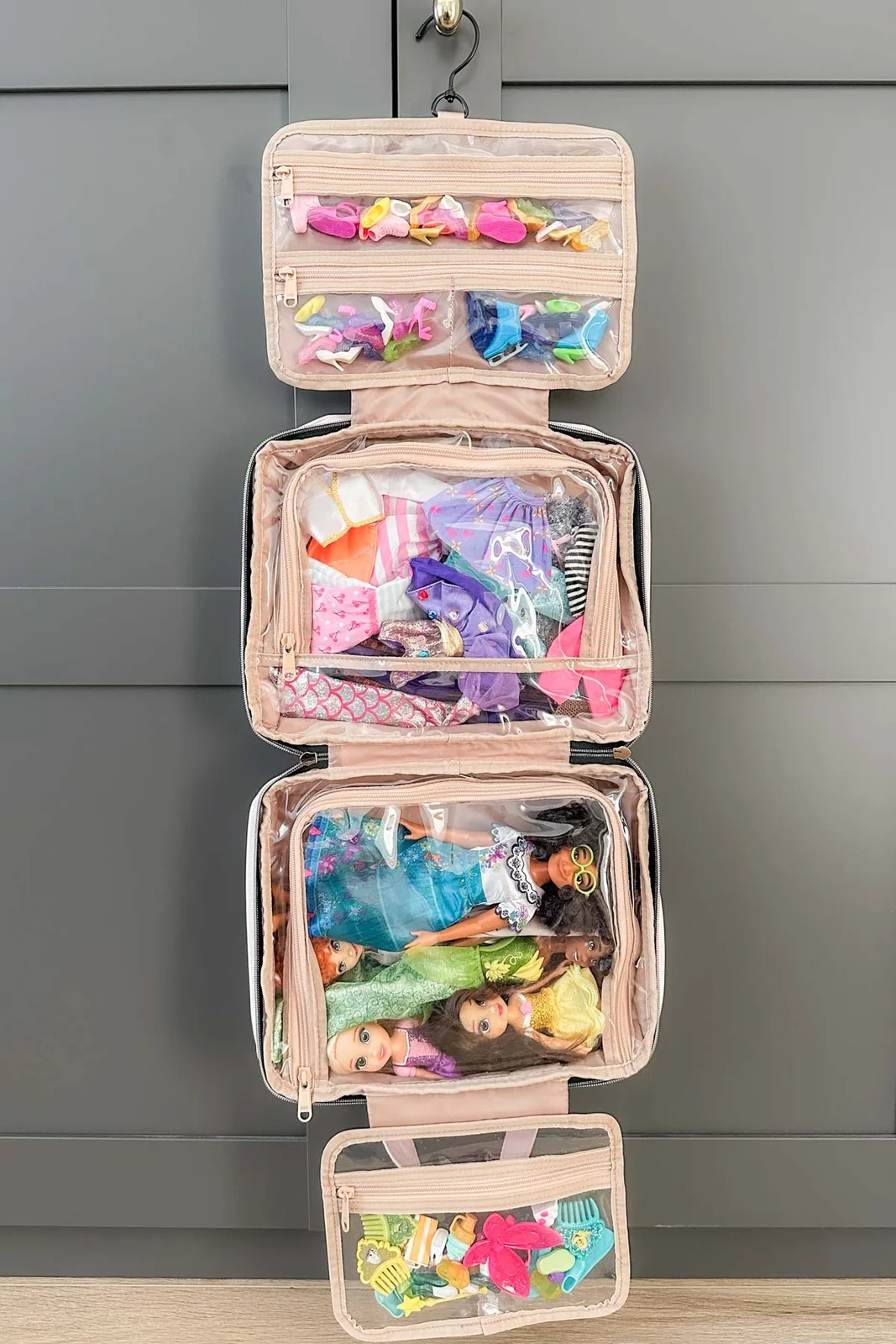 Open pink hanging travel case with clear zipped sections holding dolls, clothes, shoes and miniature brushes.