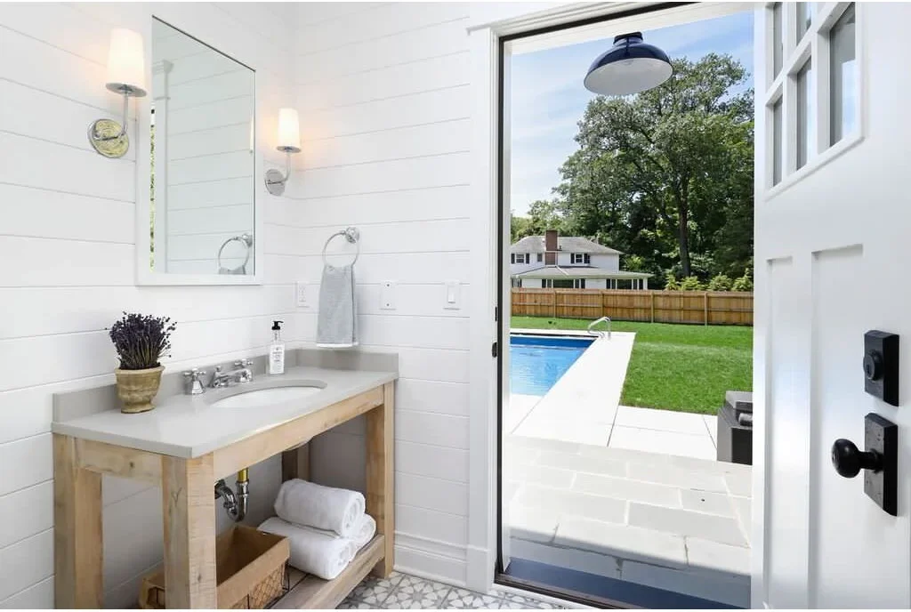 A pale towel hangs from a chrome ring beside a rectangular mirror above a wood-framed vanity and white counter.