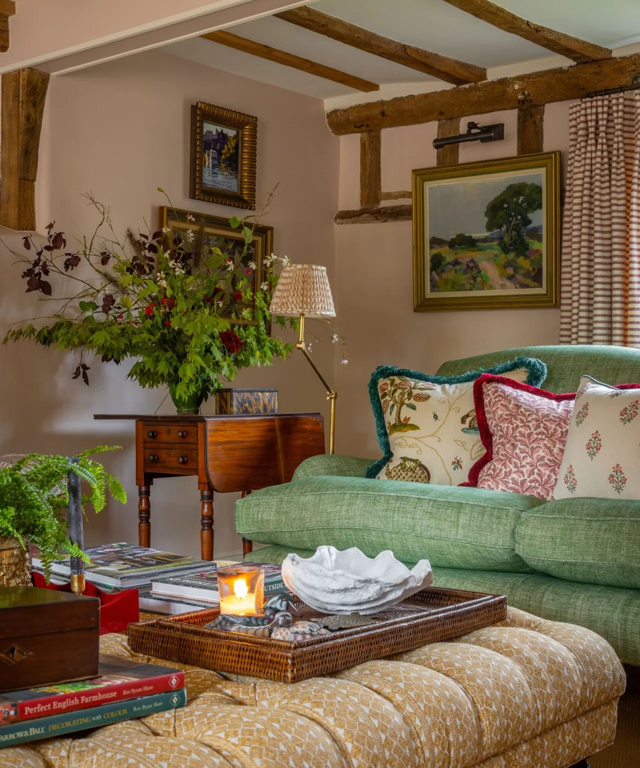 A soft green sofa with botanical cushions sits beneath wooden beams beside a wooden side table and leafy arrangement