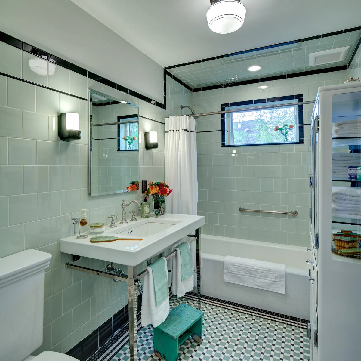 Pale green wall tile with dark borders surrounds a console sink, white bath and small mosaic floor in a vintage-style bathroom.