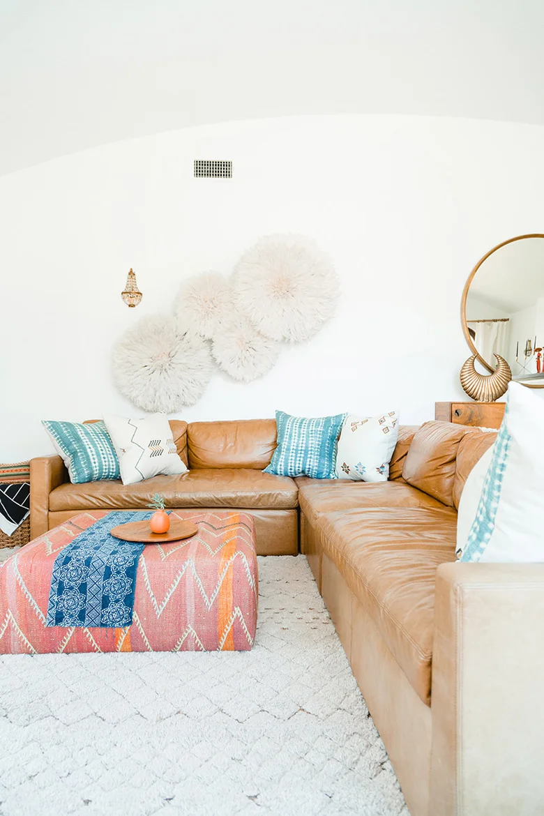 A tan sectional sits against white walls with turquoise patterned cushions, a colorful fabric-covered ottoman, pale rug and soft circular wall decorations.