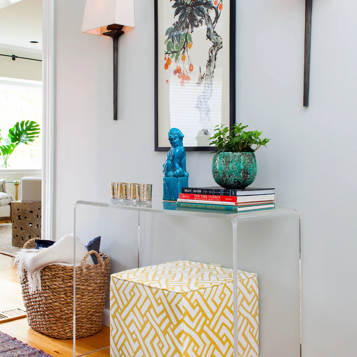 A clear console with a yellow patterned stool, books, a green planter and a blue decorative figure.