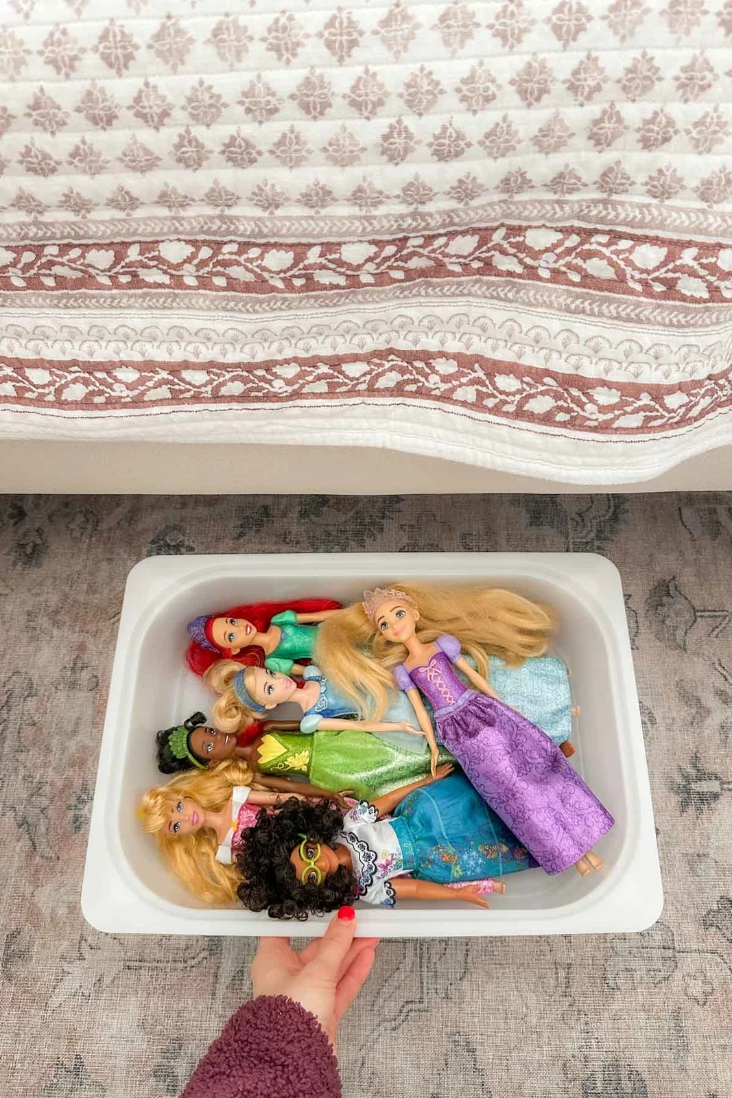 White shallow bin of dressed fashion dolls being pulled out beside a bed with patterned bedding.